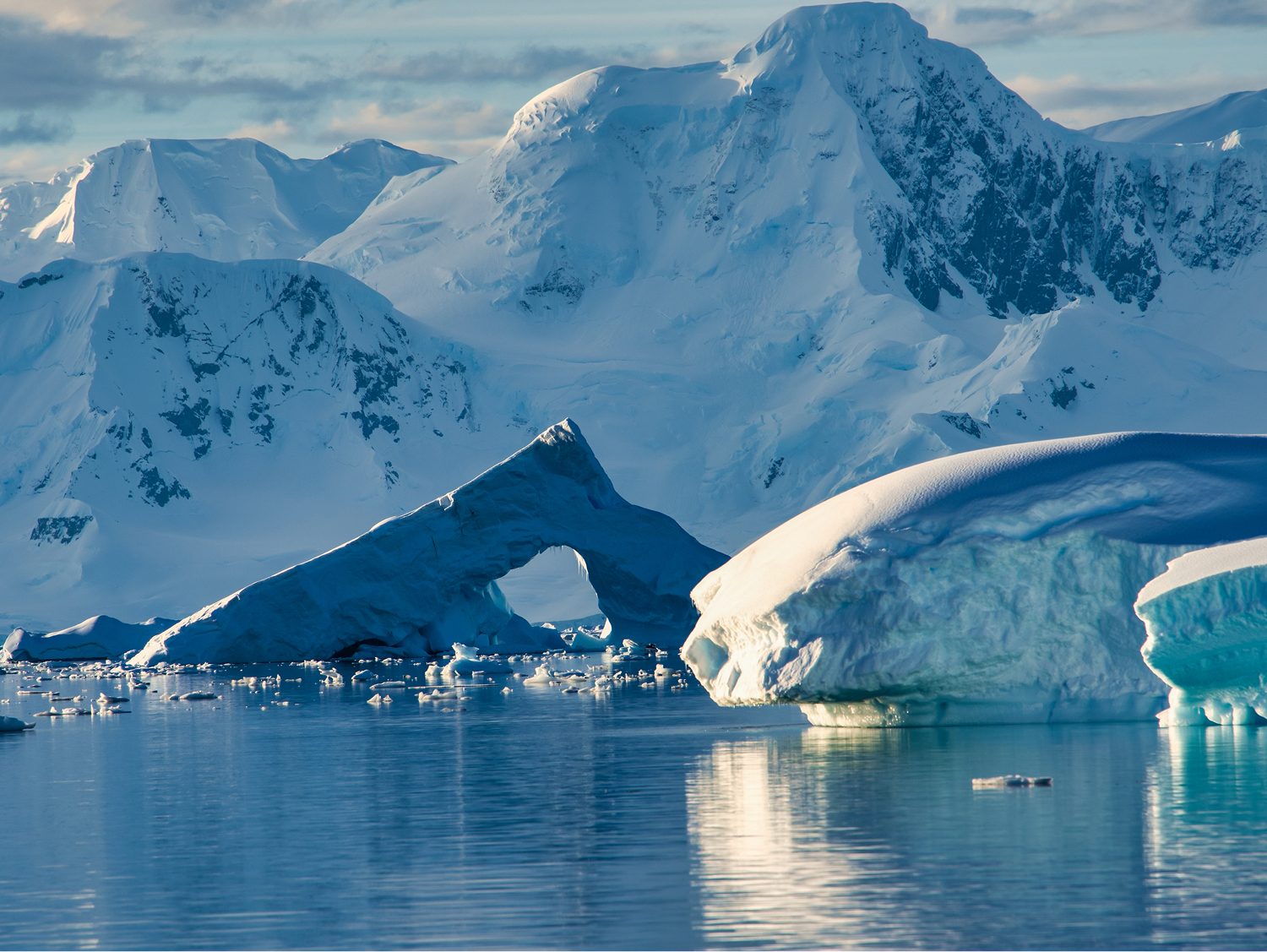Snow-covered mountains and a sculpted iceberg reflected in still water on the Antarctic Peninsula