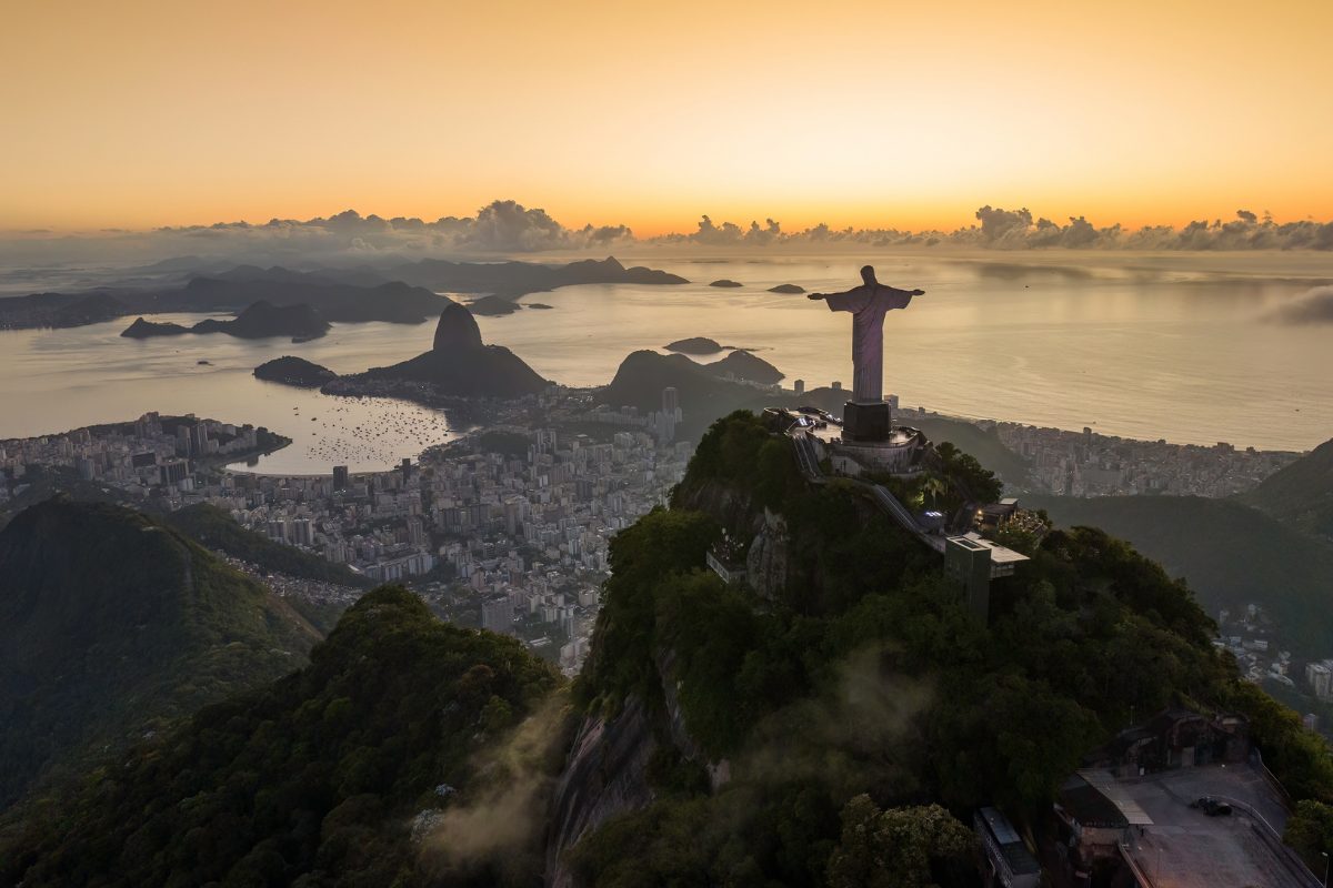 Aerial view of the Christ the Redeemer statue on Corcovado Mountain overlooking Rio de Janeiro at sunrise, with Sugarloaf Mountain and the coast in the background.