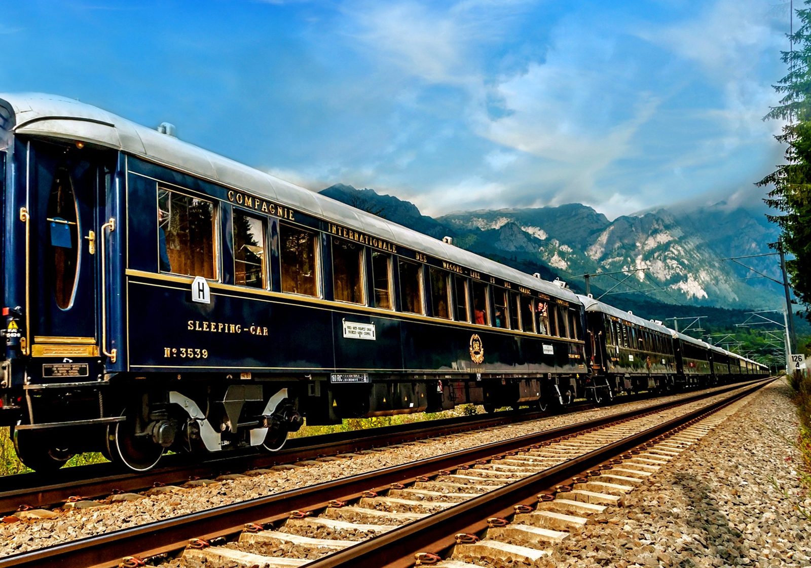The blue and gold carriages of the Venice Simplon-Orient-Express on the tracks beneath alpine mountains